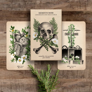 The oracle deck and two cards (hearty and cemetery) against a wooden background with rosemary sprigs below them